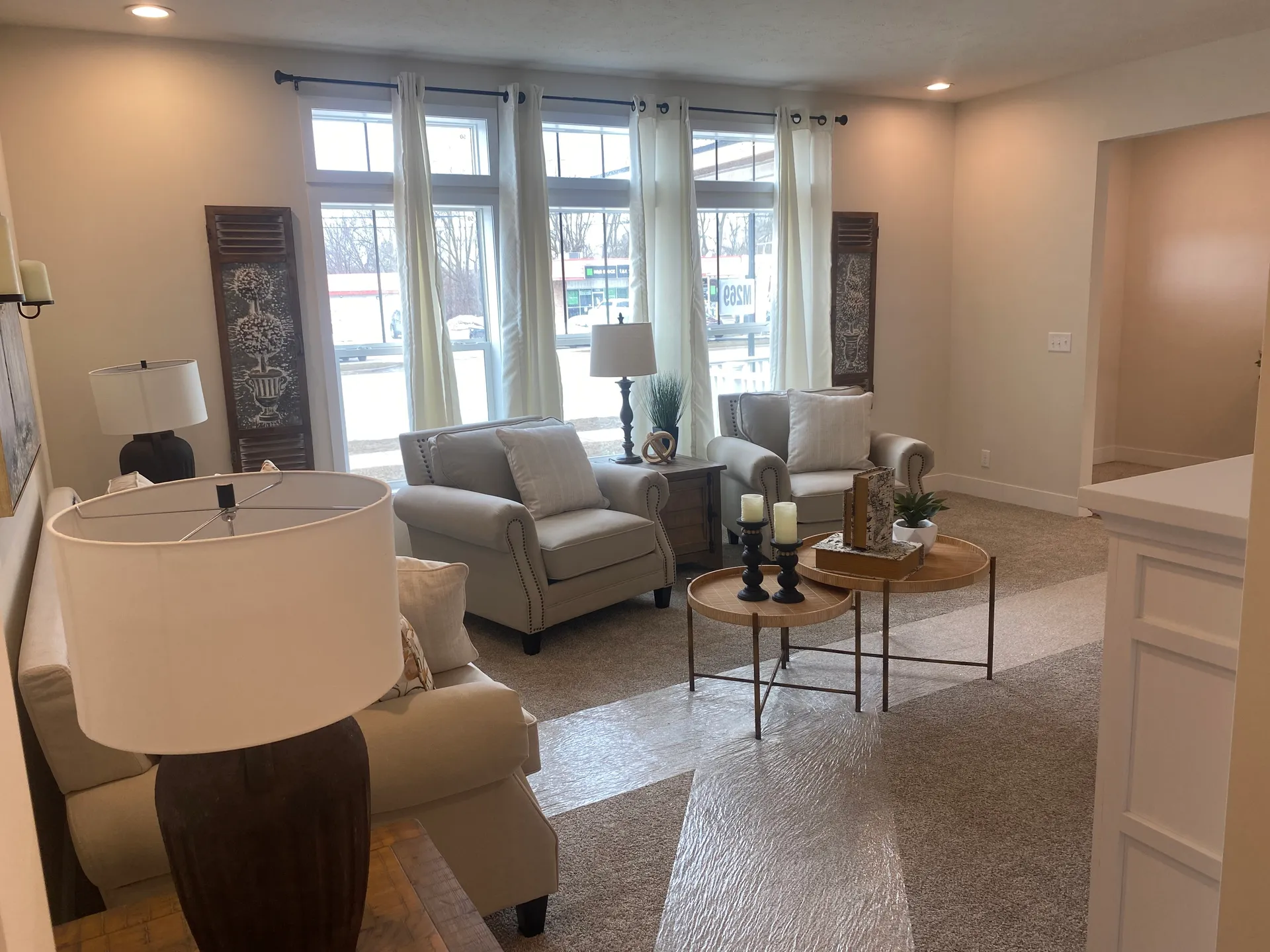 Cozy living room with beige sofas and large windows. A round coffee table with candles and decor sits on a textured rug. Soft lighting creates a warm ambiance.