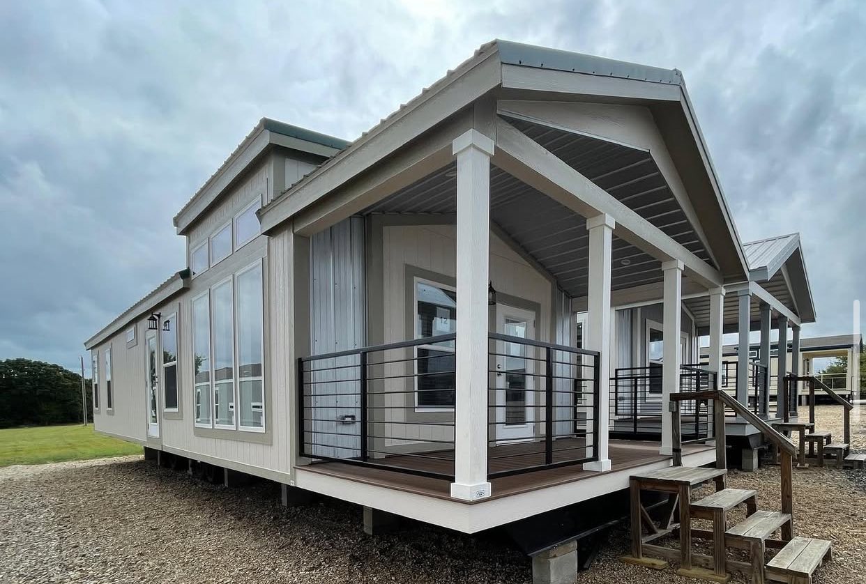 Modern tiny home with large windows and a covered porch. The light gray exterior contrasts with a cloudy sky, creating a cozy, serene atmosphere.