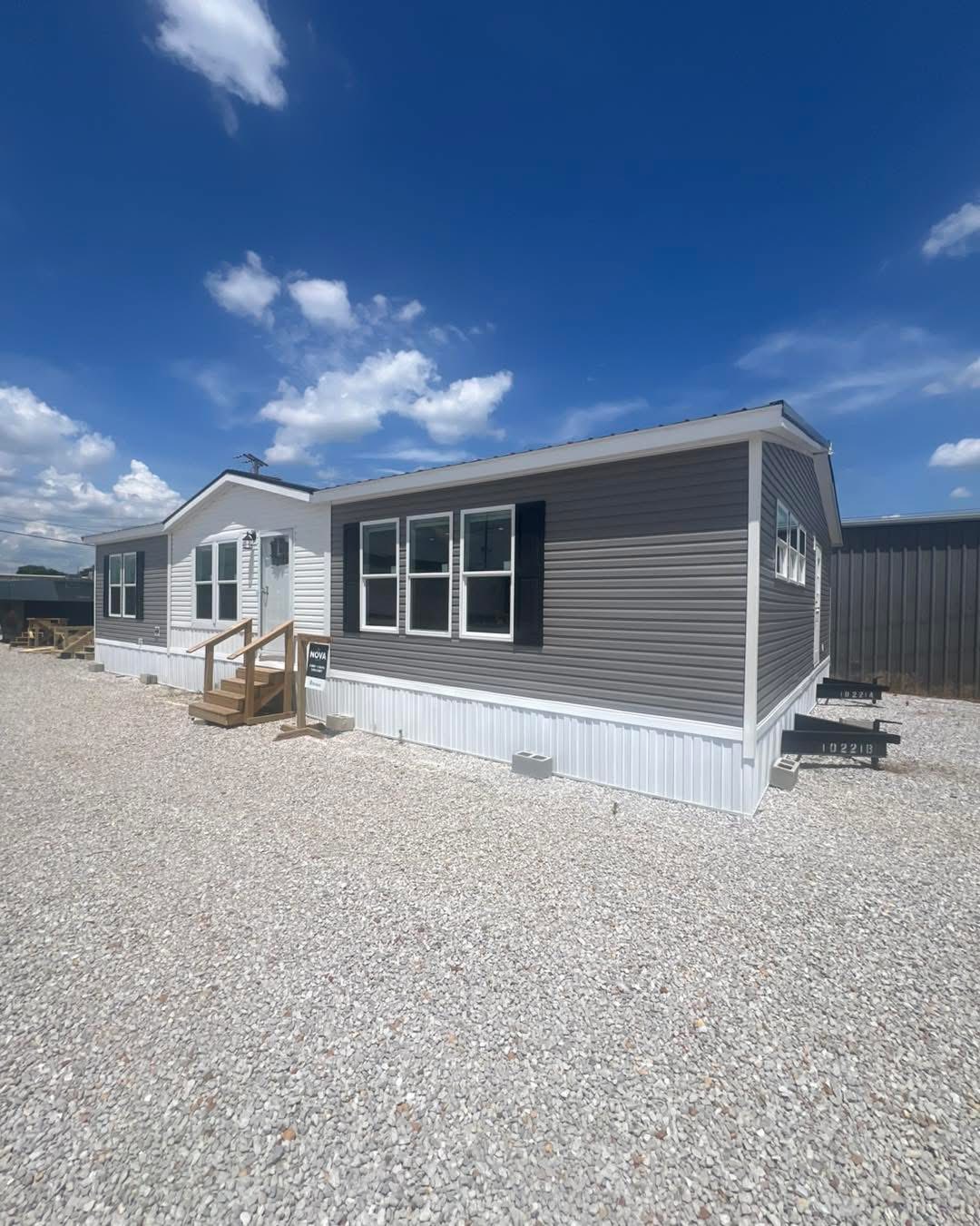 A modern mobile home with gray siding and white trim sits on a gravel lot under a clear blue sky. Wooden steps lead to the entrance, giving a welcoming feel.