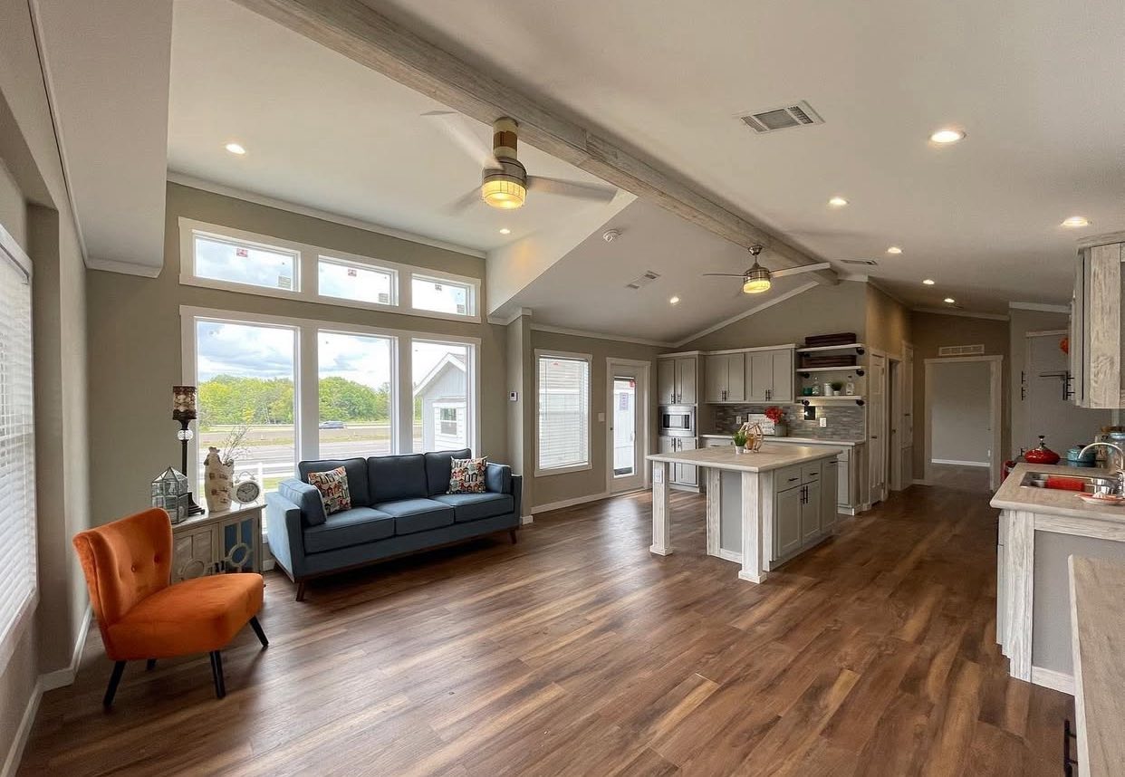 Spacious living room with a wooden floor, blue sofa, and orange chair. The open kitchen features an island and modern appliances. Bright windows offer a scenic view.