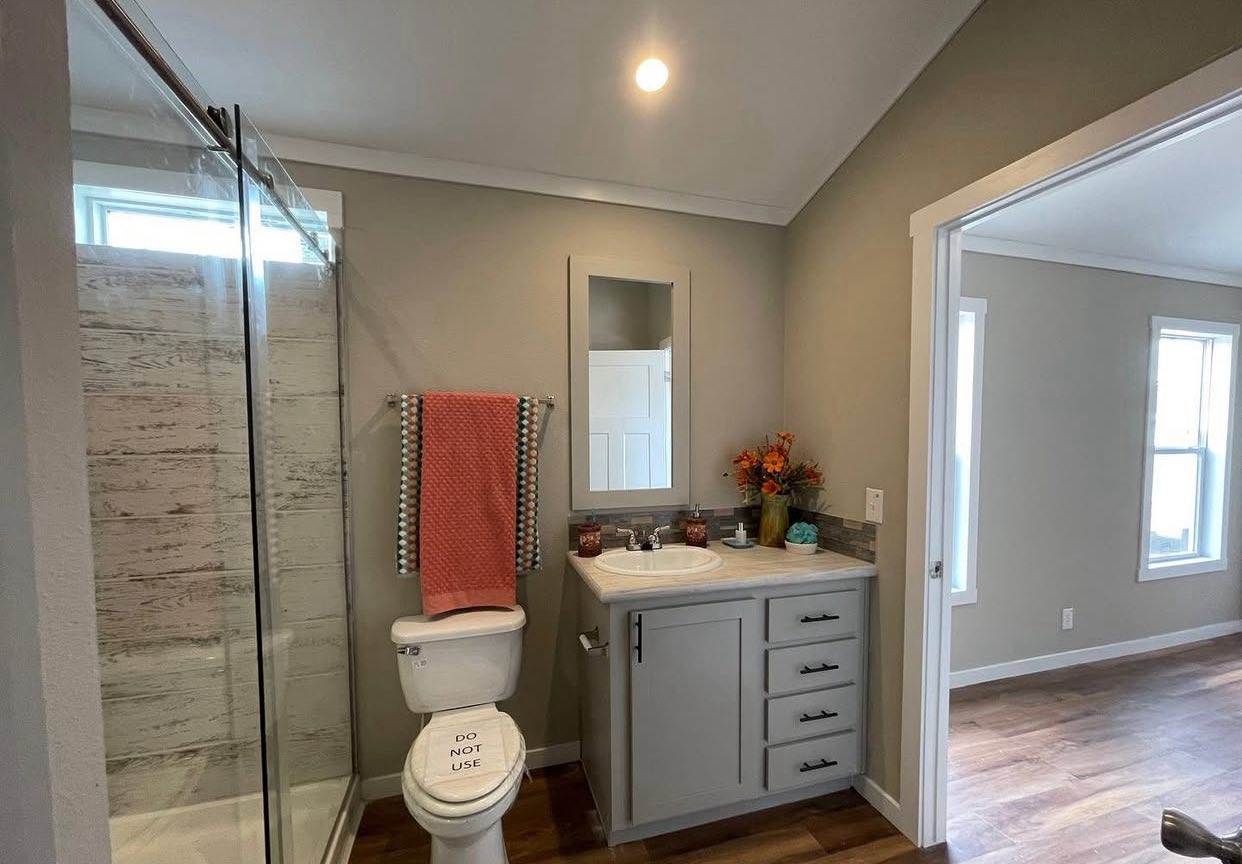Bright bathroom with glass shower, gray vanity, and mirror. A colorful towel hangs above a toilet labeled “Do Not Use.” Flowers add a lively touch.