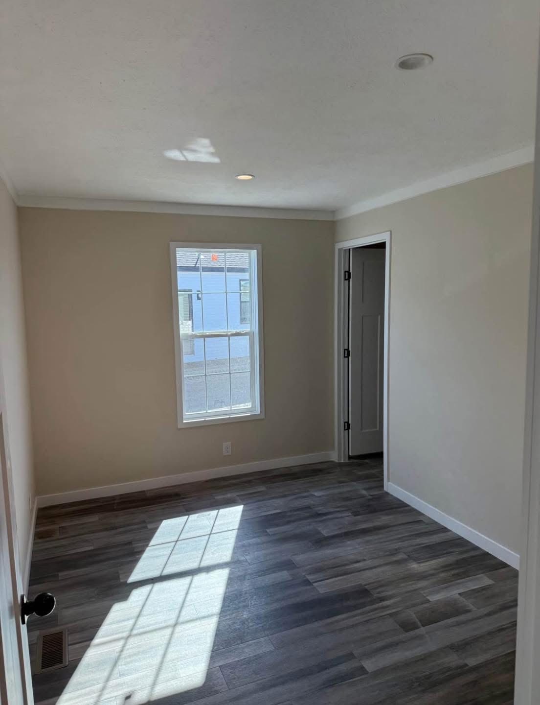 A small, empty room with beige walls and gray wood flooring. Sunlight streams through a single window, casting shadows. The room is minimalist and peaceful.