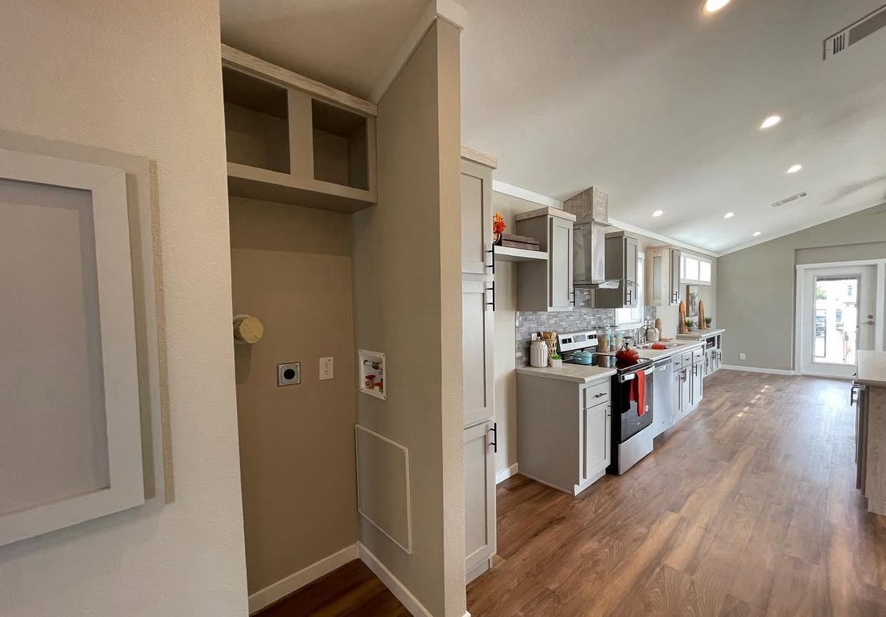 Modern kitchen with light gray cabinets, stainless steel appliances, and a long wooden floor. Bright lighting gives a cozy, inviting feel.