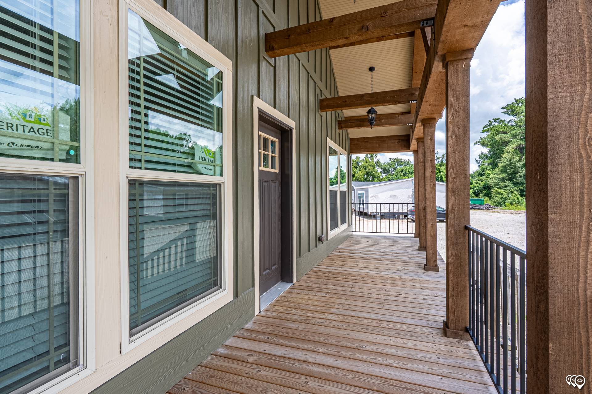 A wooden porch with a rustic design features large windows, a dark wooden door, and sturdy beams. Bright and inviting, it overlooks a gravel driveway.