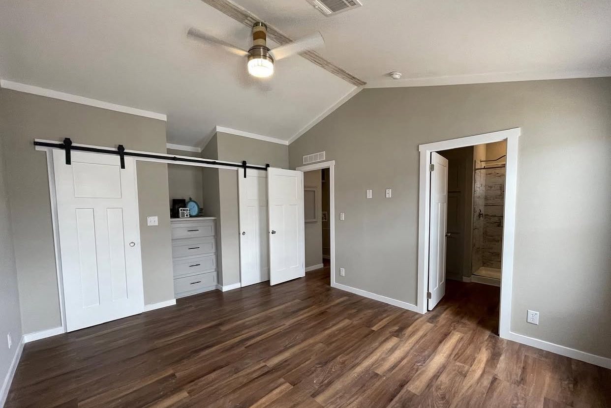 A spacious, minimalist bedroom with light gray walls and wood flooring. It features double sliding closet doors, a ceiling fan, and a door to an adjoining bathroom.