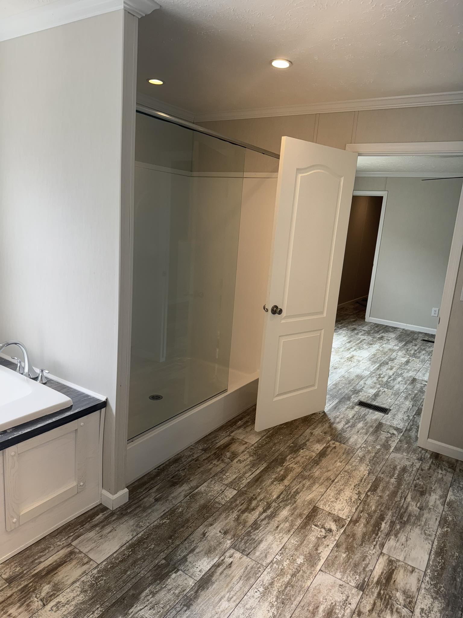 Bathroom with wood-textured tile flooring, glass shower on the left, partially open door leading to a bright hallway. Calm, clean, modern vibe.