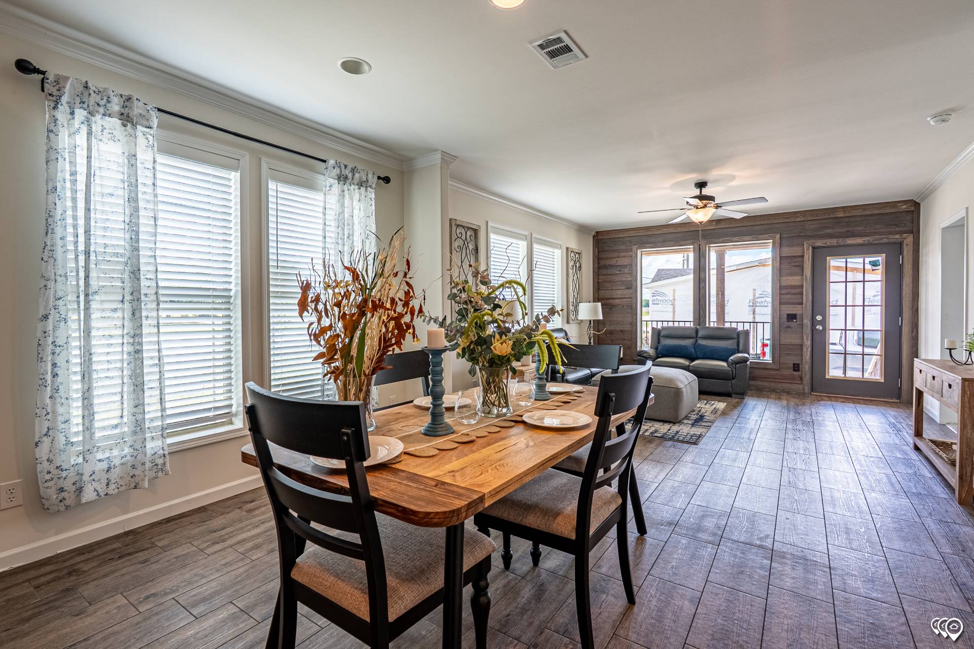A cozy living-dining area with wooden flooring, large windows, and white curtains. A wooden dining table with chairs is set with plates and flowers. A sofa and fan are seen in the background.