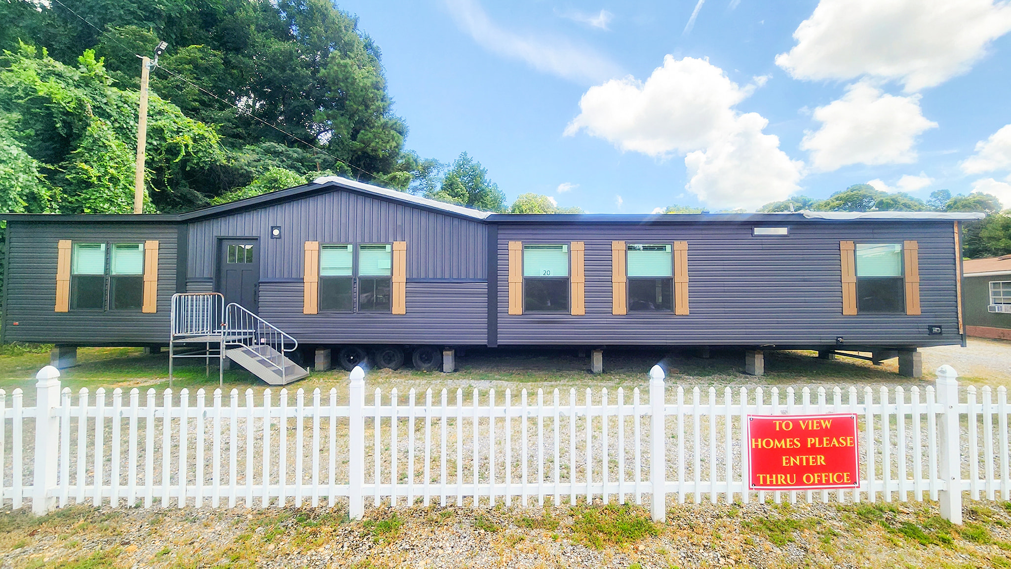 A modern, dark gray manufactured home stands behind a white picket fence, with lush green trees in the background and a sign directing visitors through the office.