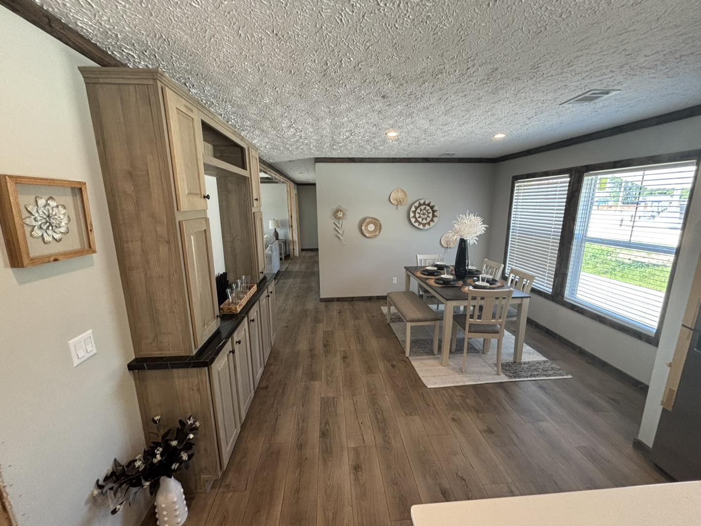 Bright dining room with wooden floors, beige cabinets on the left, and modern decor. A table is set for four by large windows, creating a cozy ambiance.