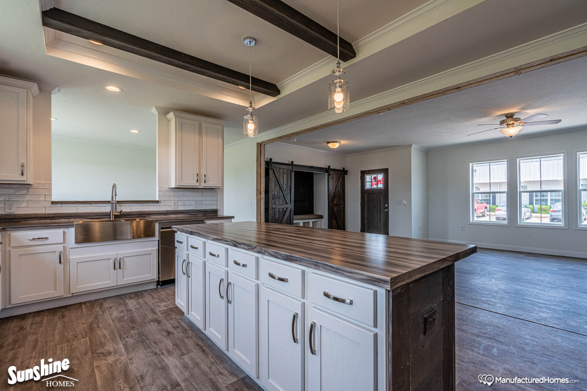 Spacious kitchen with white cabinets, wood island, and beams. Stainless steel sink, pendant lights, and adjacent living room with barn doors. Airy and modern.