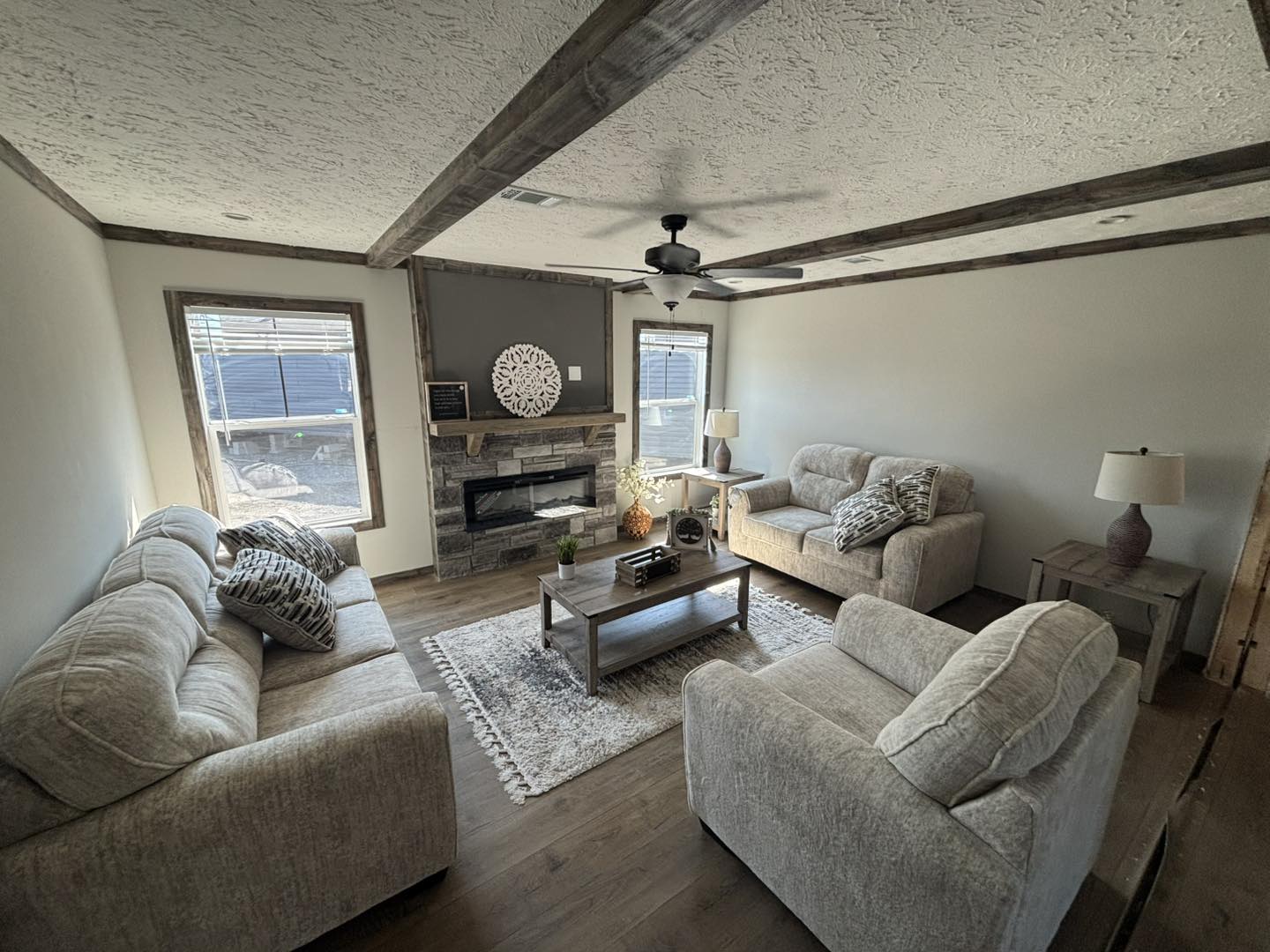 Cozy living room with beige couches, a wooden coffee table, a stone fireplace, and rustic beams. Sunlight streams through windows, creating a warm ambiance.