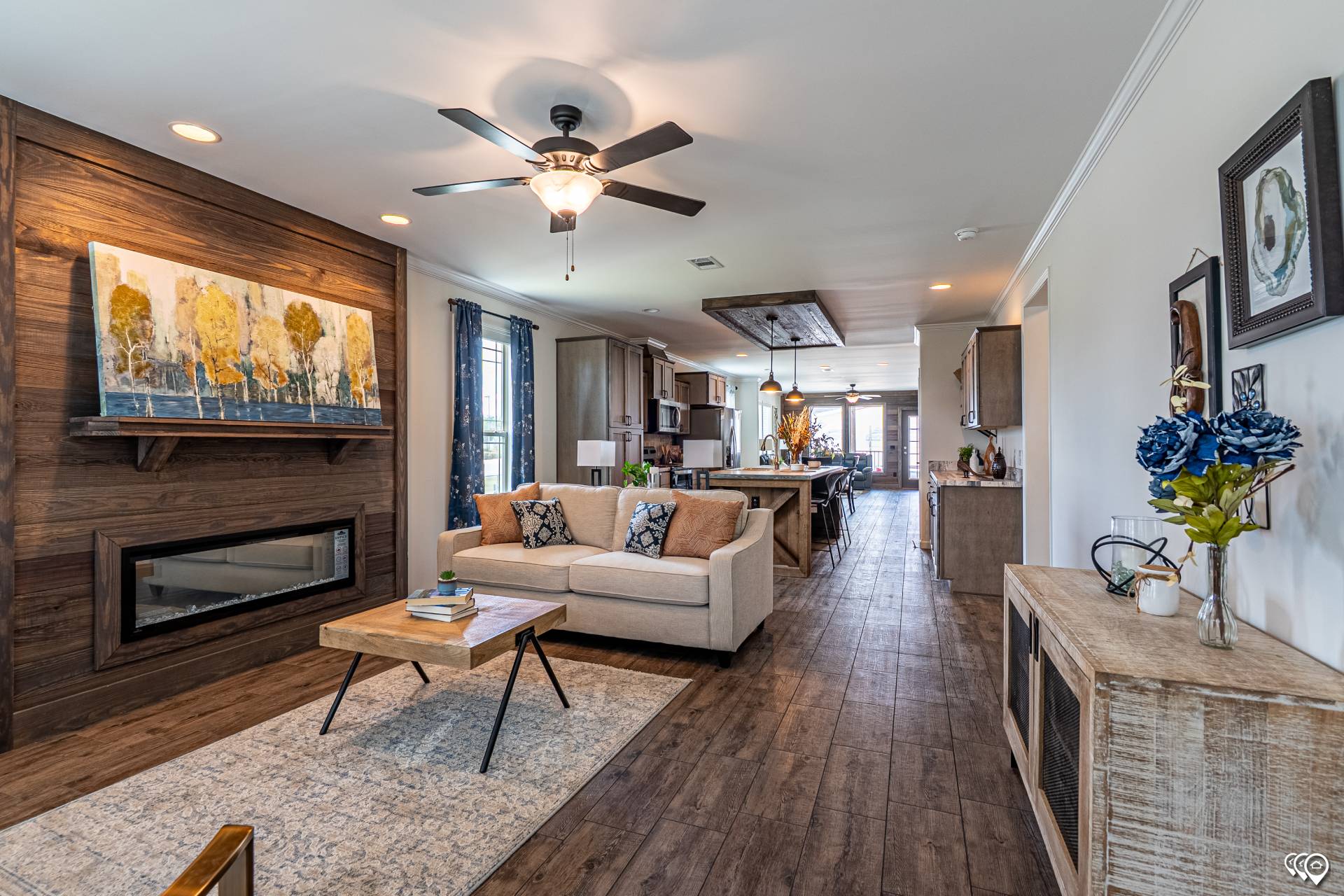 Stylish open-concept living room and kitchen. Features a beige couch, wood floors, decorative fireplace, ceiling fan, and large artwork. Bright and inviting.
