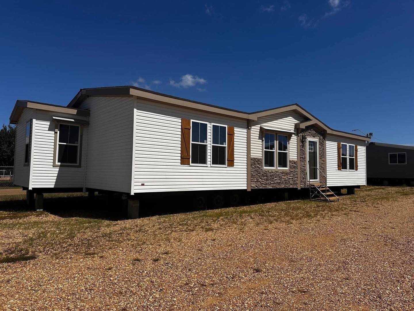 A white modular home with brown shutters and stone accents sits on a gravel lot under a clear blue sky, conveying a sense of tranquility and simplicity.