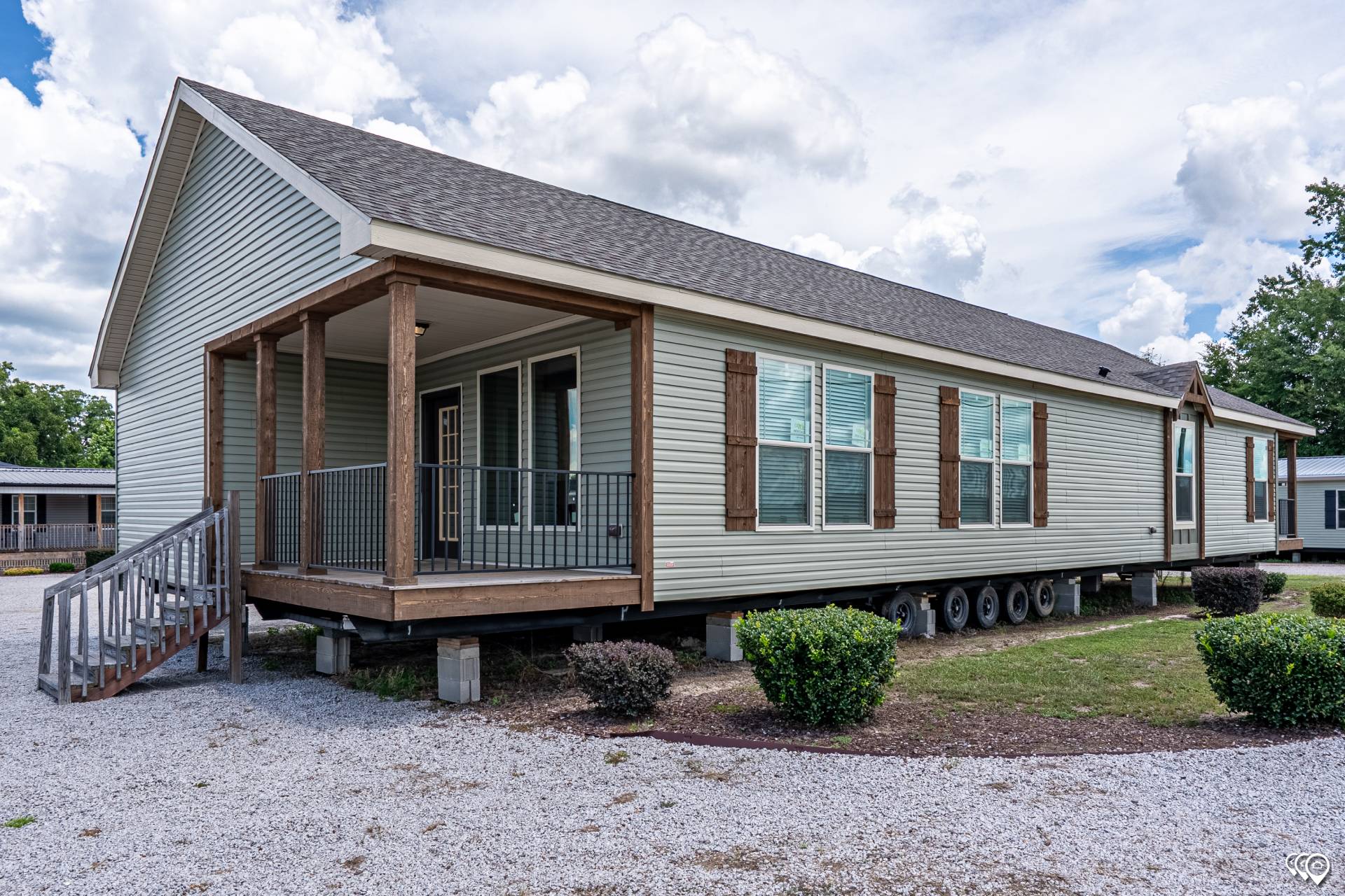 A mobile home on a gravel lot, featuring light siding, wooden shutters, a small porch with wooden railings, and stairs. The sky is partly cloudy.