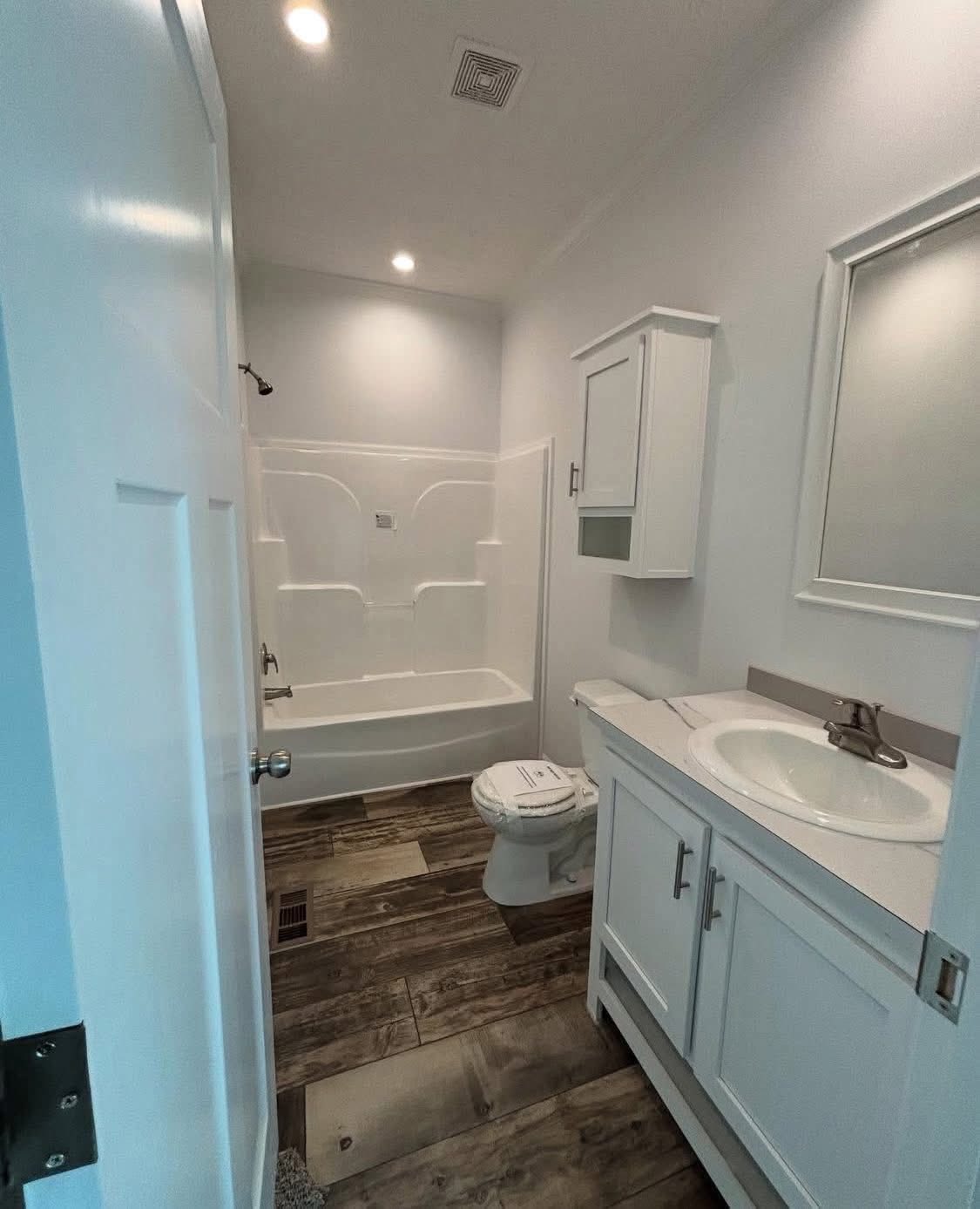 Small bathroom with a wooden floor, white walls, and ceiling lights. There's a bathtub at the back, a toilet beside it, and a sink with a mirror.
