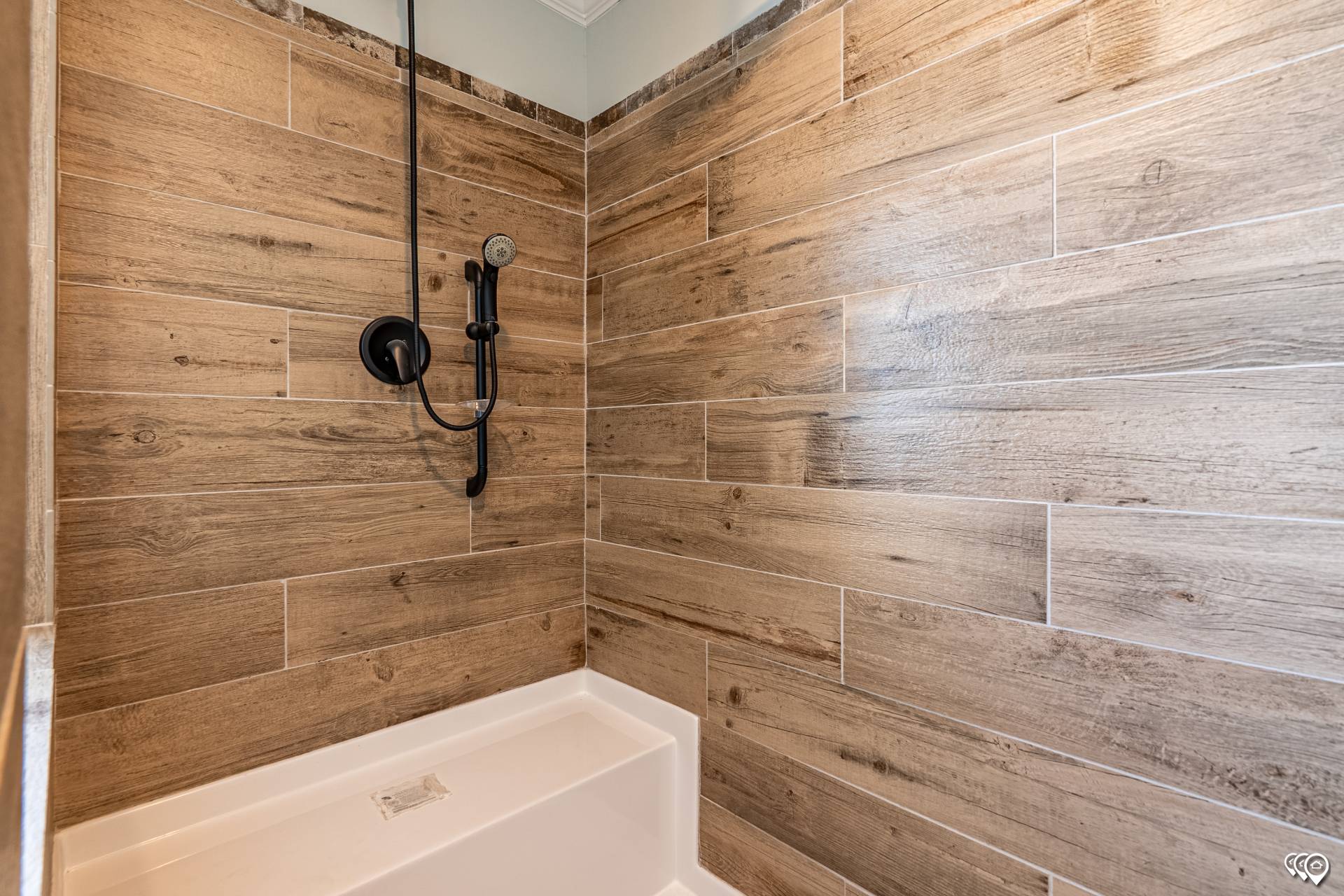 Shower with wood-look tiled walls and a black showerhead, creating a rustic and modern feel. Bright lighting highlights the textured finish.