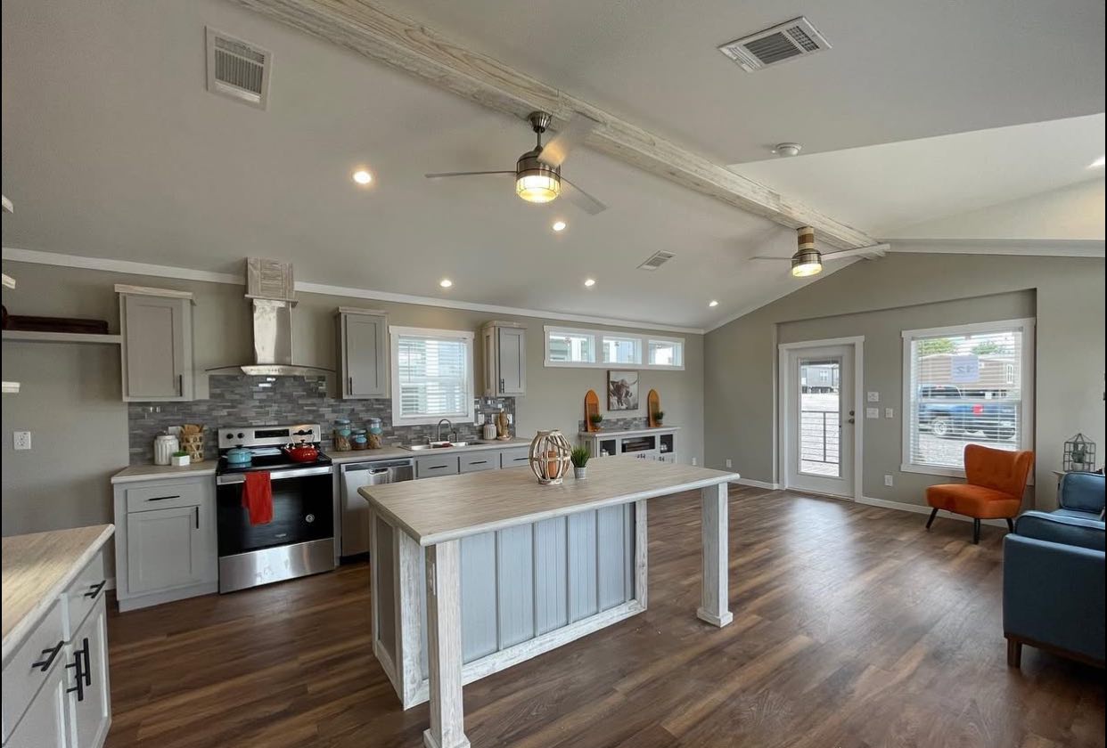 Spacious modern kitchen with light wood floors, gray cabinets, and a central island. Bright windows, orange chair, and ceiling fans create an airy feel.