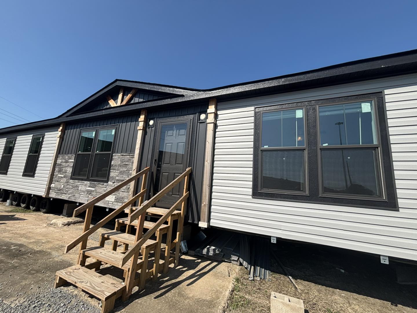 Single-story modular home with gray siding and stone accents under a clear blue sky. Wooden stairs lead to a dark front door, conveying a welcoming tone.