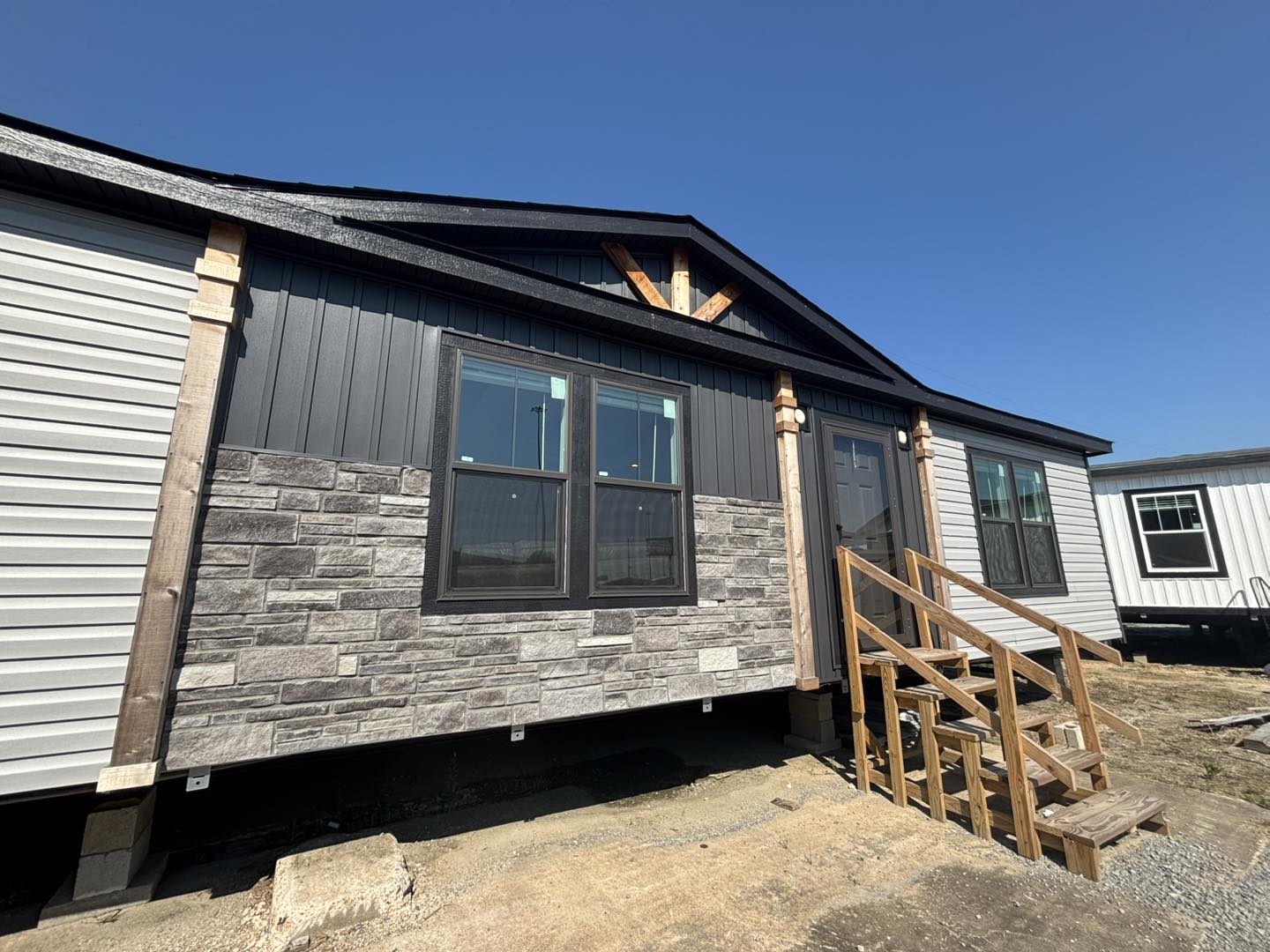 Modern manufactured home with stone and wood siding, two large windows, and wooden steps leading to the entrance, set against a clear blue sky.
