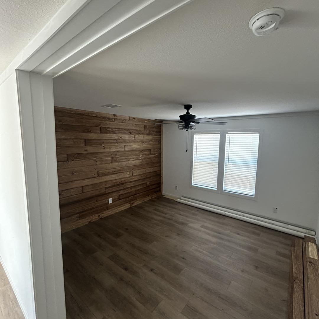 An empty room with wood panel flooring and a wood accent wall, featuring a ceiling fan and two windows with blinds. The atmosphere is clean and minimalist.