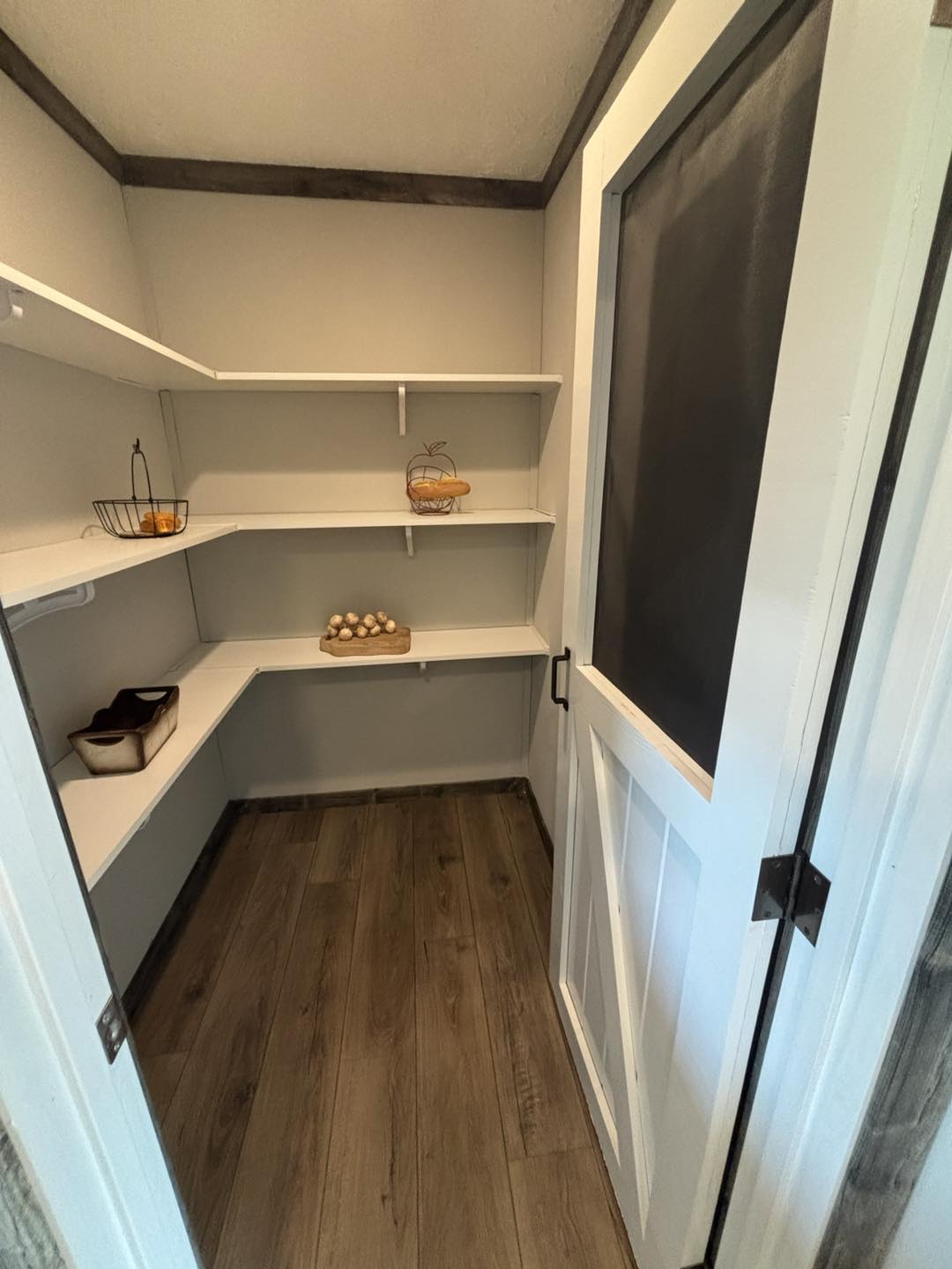 Small pantry with white shelves and wood flooring. Shelves hold a wire basket with oranges, a decorative item, and eggs. Door partially open.