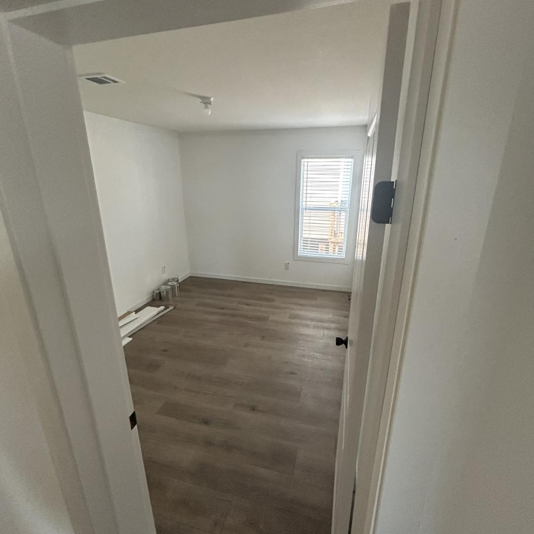 Empty room with light wood flooring and white walls, viewed through an open door. A window with blinds allows sunlight to fill the space. Construction materials sit in the corner.