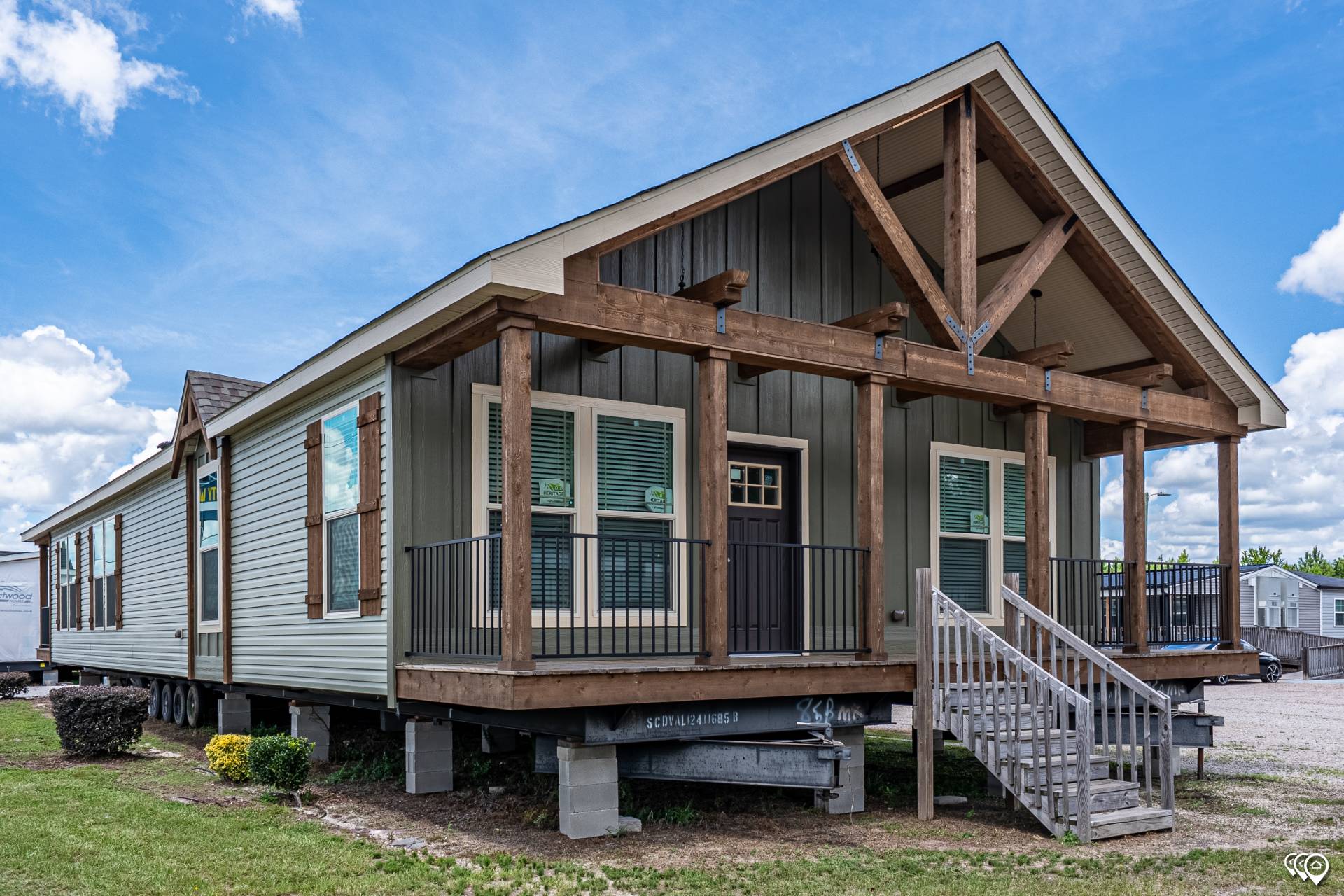 A modern modular home with a wooden porch and steps, featuring large windows and a peaked roof. The setting is sunny, with fluffy clouds in a vivid blue sky.