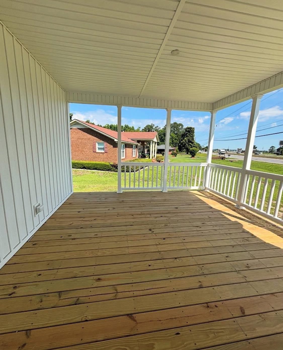 Spacious wooden porch with white railing and walls, overlooking a grassy yard and a brick house under a clear blue sky, conveying a serene atmosphere.