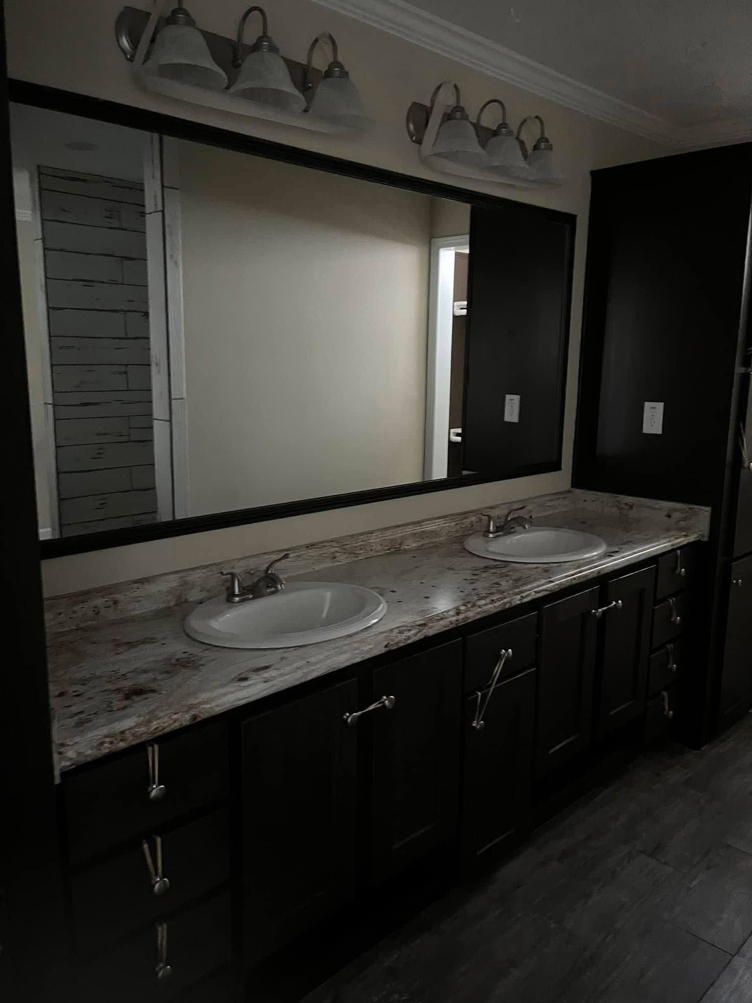 Dark bathroom with two sinks on a granite countertop, large mirror, and dual overhead light fixtures. Dim lighting creates a somber tone.