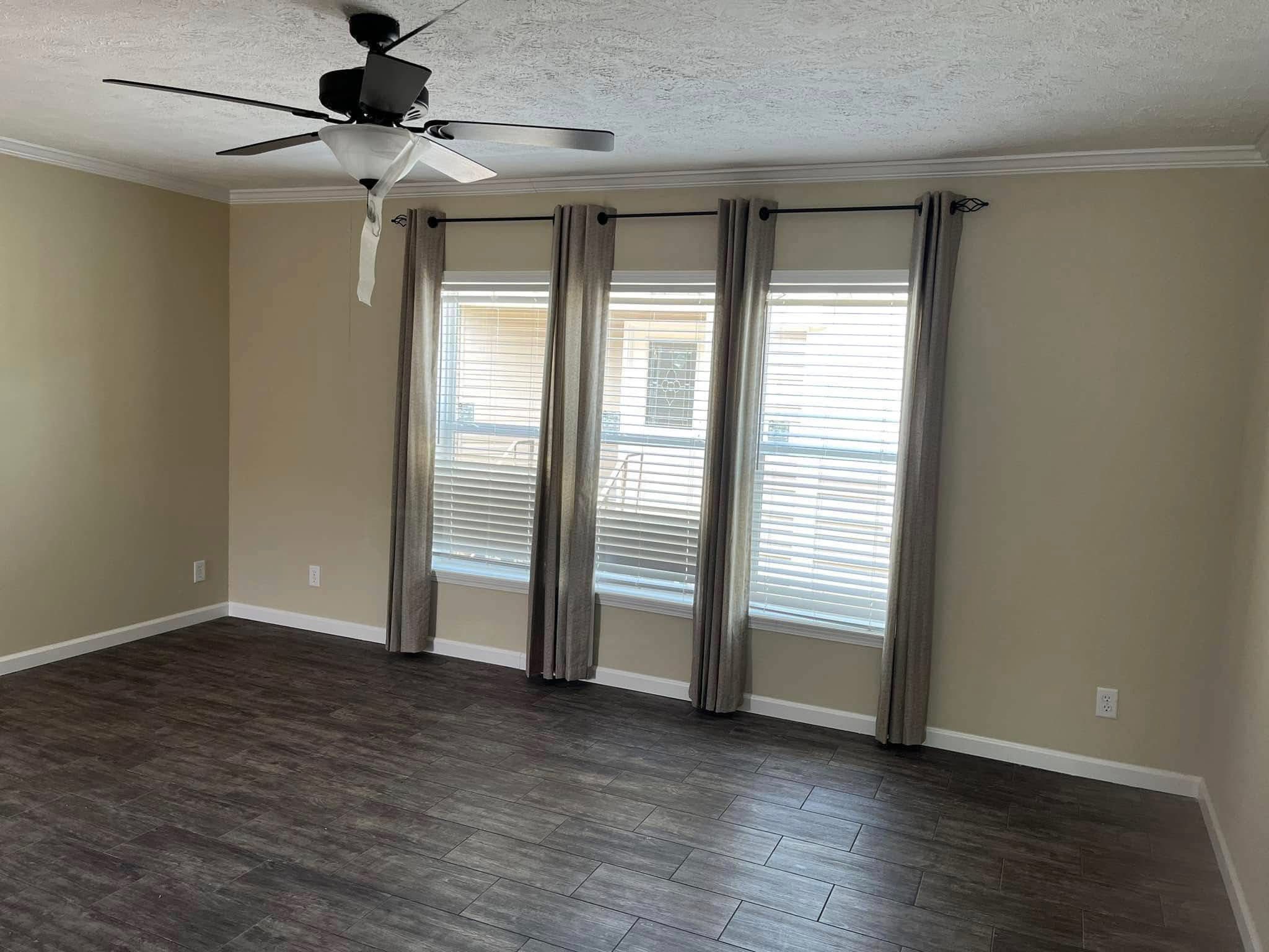 Empty room with dark wood floors, beige walls, and three windows with gray curtains. A ceiling fan hangs from a white ceiling. Natural light filters in.