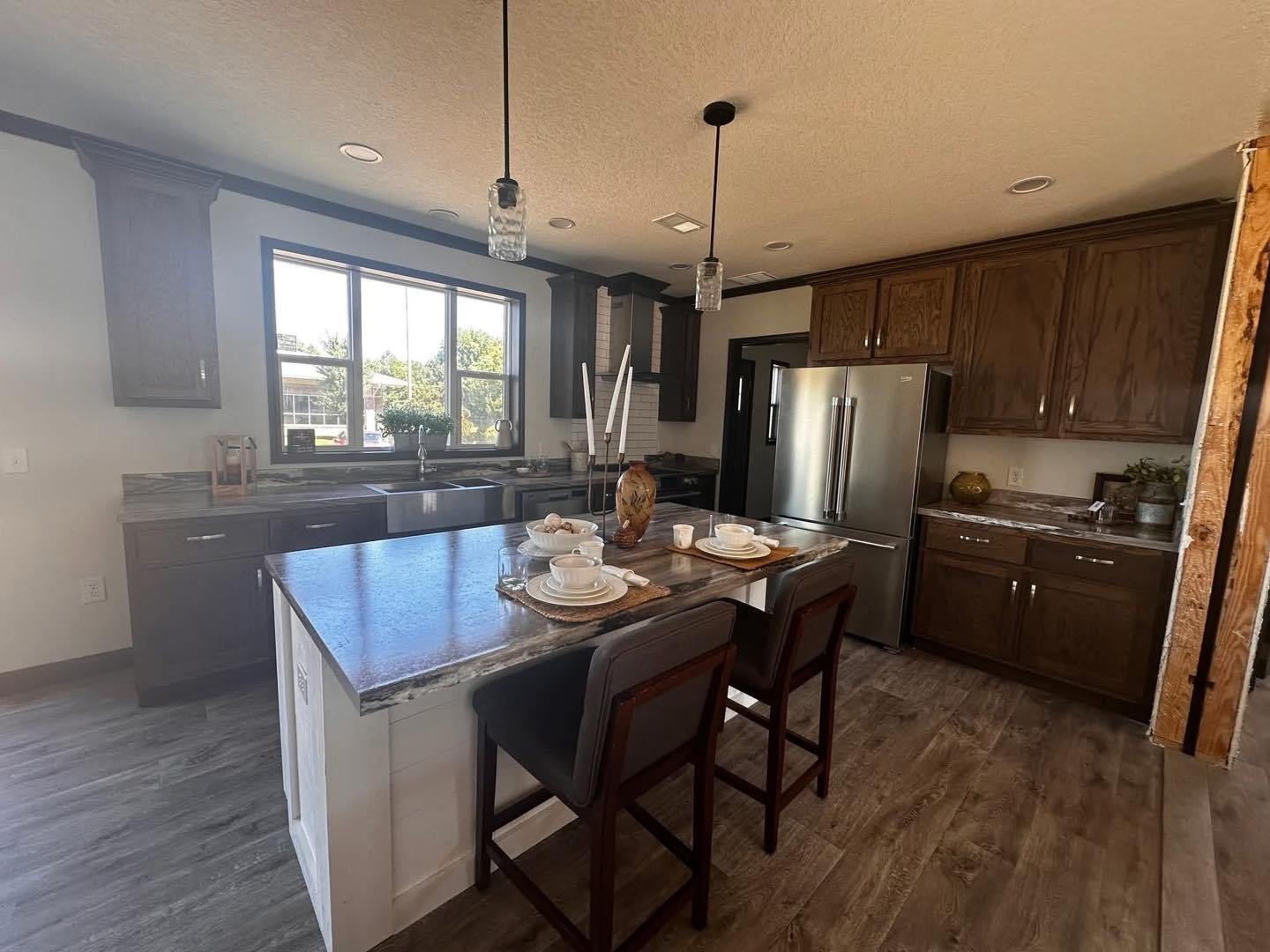 Modern kitchen with dark wood cabinets, stainless steel fridge, and marbled countertops. Island with bar stools set for dining, large window offers natural light.