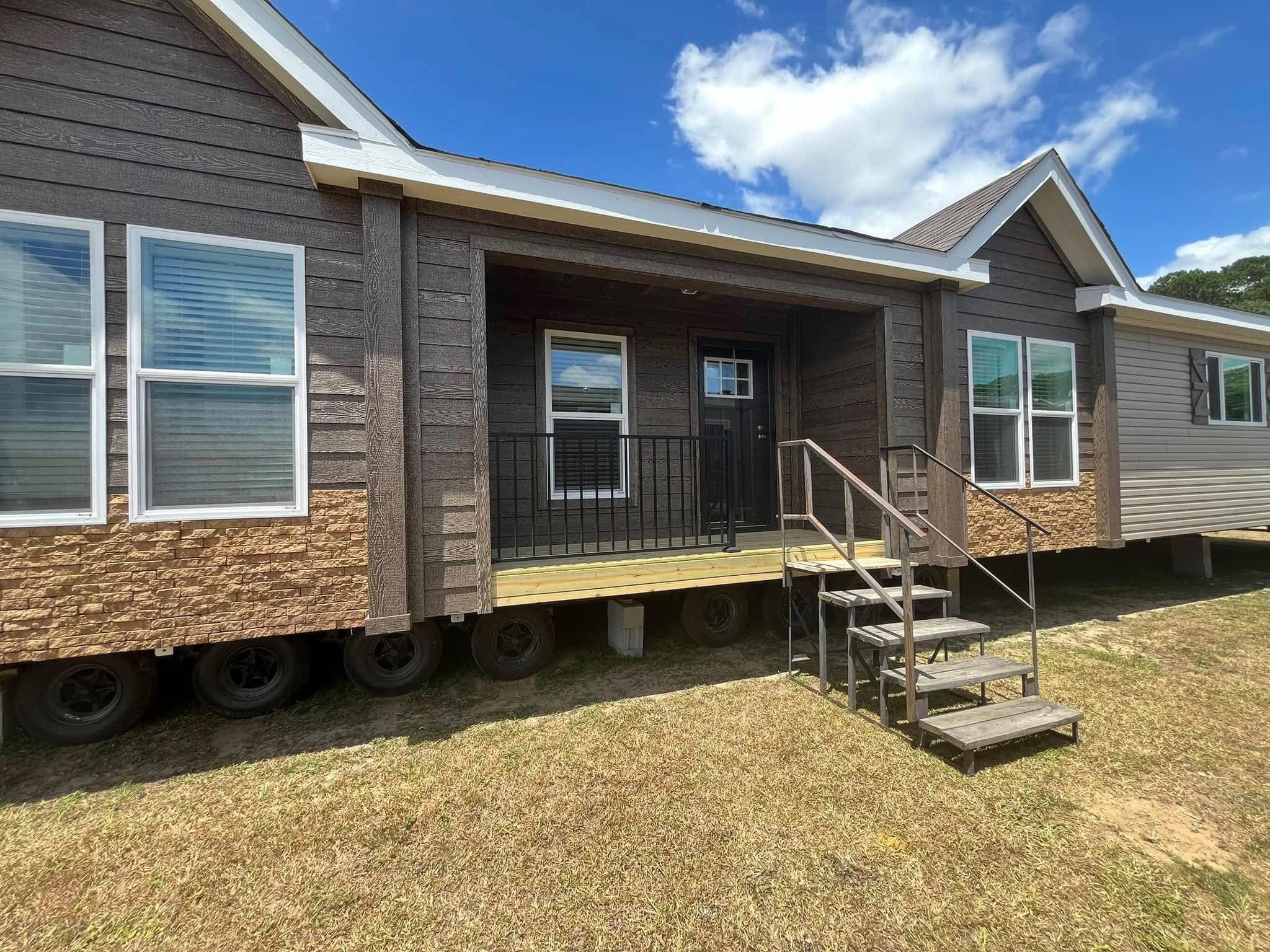 Brown modular home with a small porch and staircase. The siding is a mix of wood and stone textures. Bright blue sky with clouds in the background.