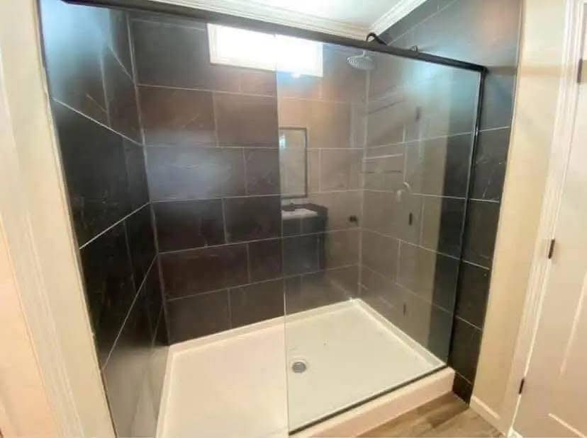 Modern bathroom with glass-enclosed shower, dark tiles, and light flooring. A small window above adds brightness. Sleek, clean design.