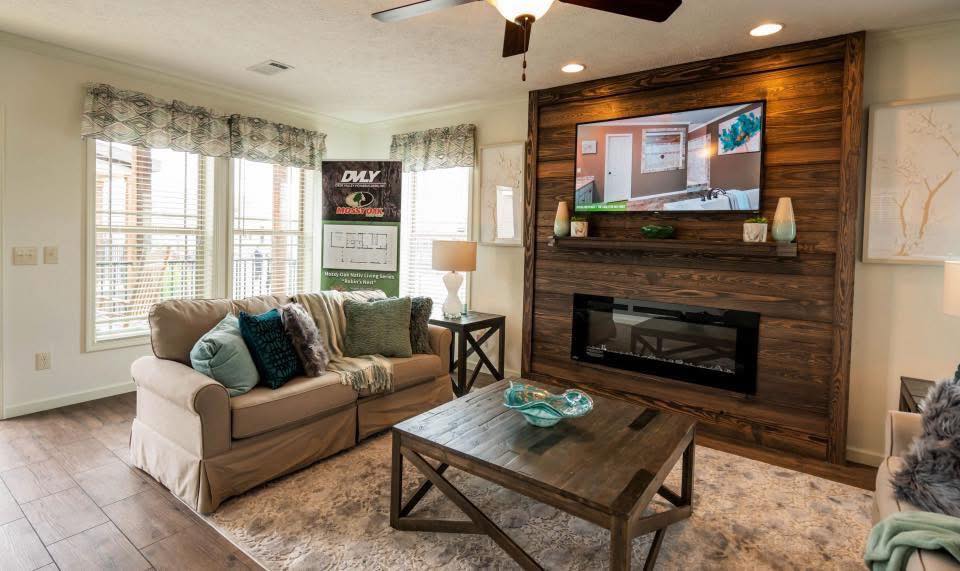 Cozy living room with a beige sofa, teal cushions, and a wooden coffee table on a plush rug. A TV is mounted above a fireplace on a wooden accent wall, creating a warm, inviting atmosphere.