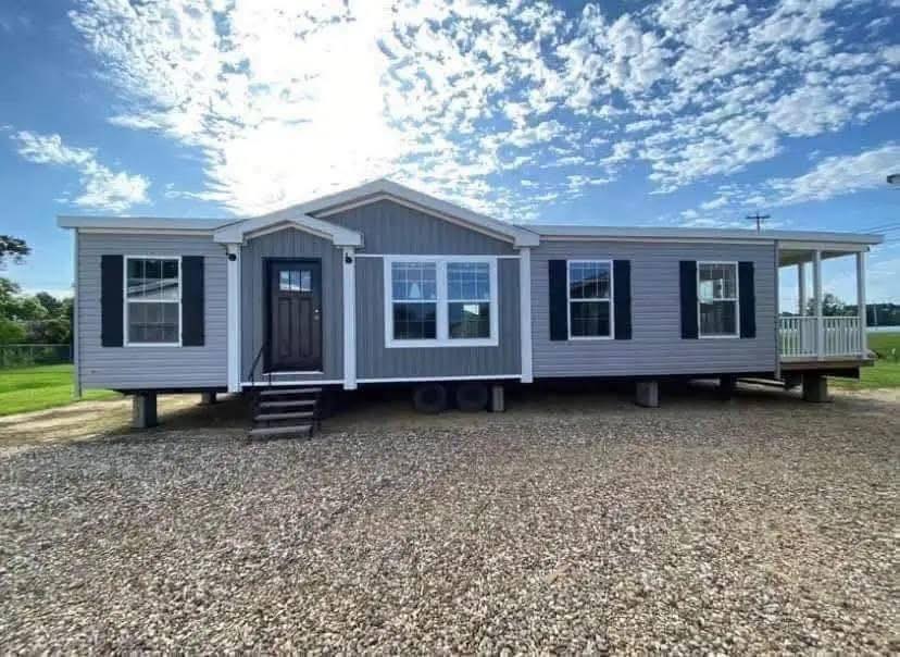 A gray modular home with a pitched roof is set against a bright sky and scattered clouds. The house has white trim, black shutters, a small porch, and a gravel driveway.
