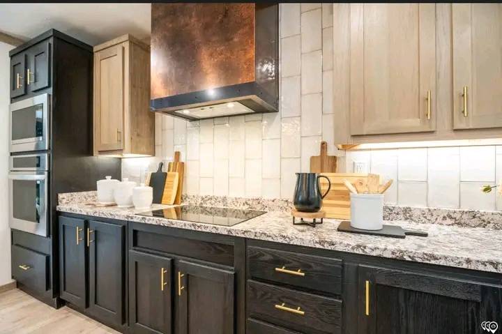 Modern kitchen with dark cabinets, brass handles, and a marble countertop. Features a copper range hood and tiled backsplash with warm lighting.
