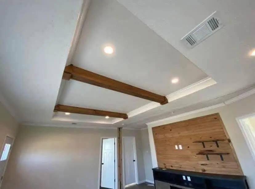 Spacious room with white walls, recessed lighting, and wooden ceiling beams. The back wall features wood paneling and floating shelves, creating a warm, modern ambiance.