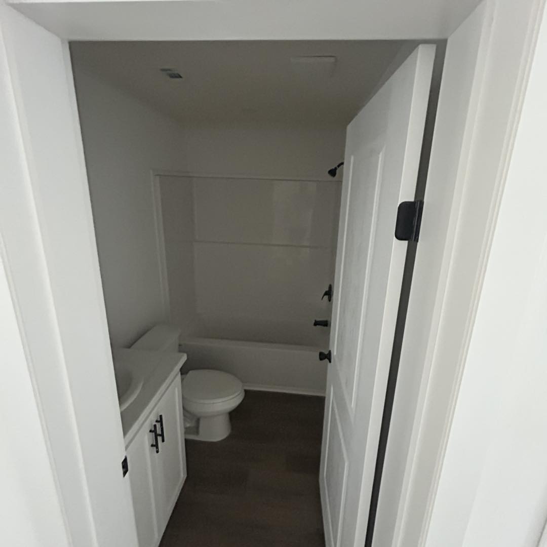 Small, minimalist bathroom with white walls and door, featuring a bathtub, toilet, and sink with cabinet. The space appears compact and clean.