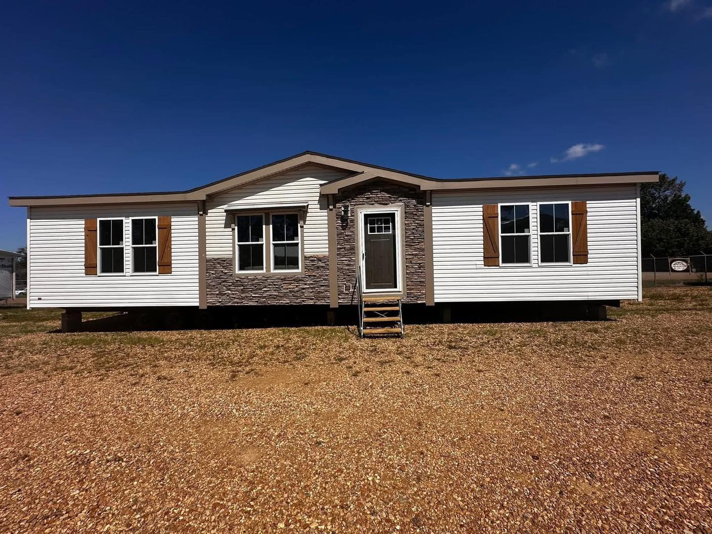 A single-story, white manufactured home with stone accents, wooden shutters, and a gray door. It sits on a gravel lot under a clear blue sky.