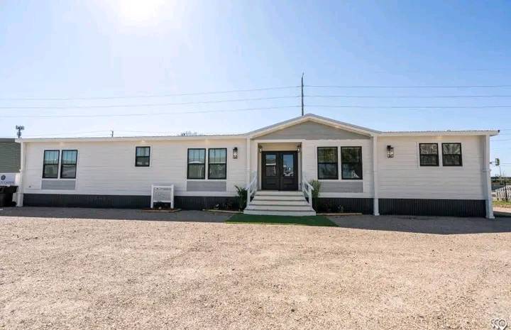 A modern, single-story manufactured home with a white facade and dark roof. The front includes steps leading to a central door, flanked by large windows.