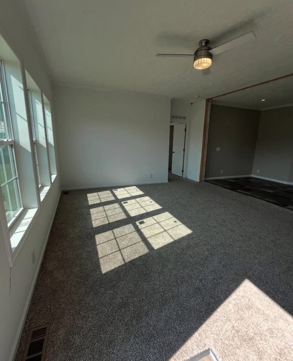 Sunlit room with beige carpeting, white walls, and a modern ceiling fan. Large windows cast symmetrical shadows on the floor, creating a bright, airy feel.