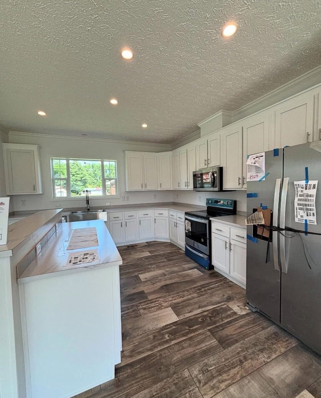 Bright, modern kitchen featuring white cabinetry, stainless steel appliances, and wood-patterned flooring. Large window and recessed lighting add openness.