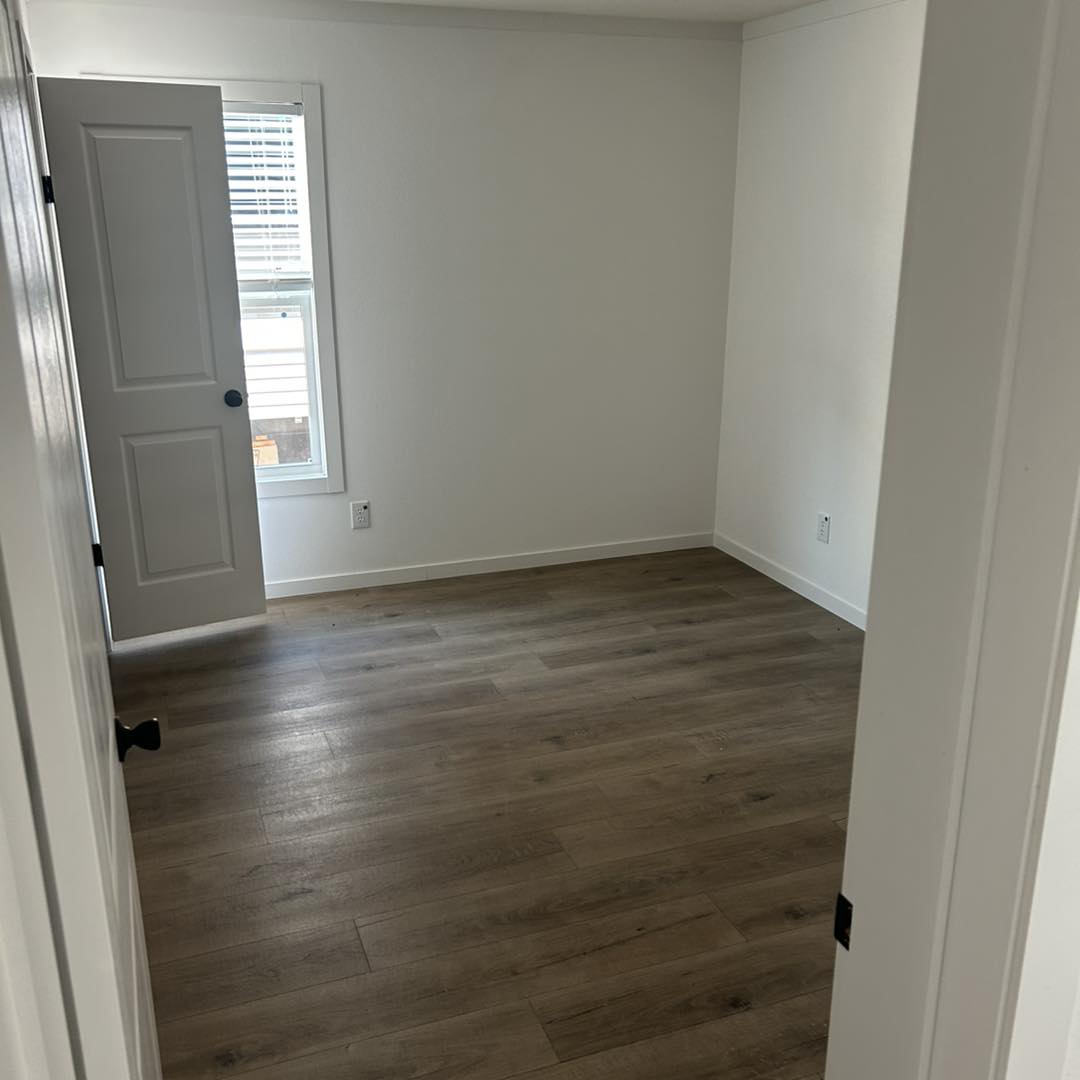 An empty, minimalist room with white walls, a wooden floor, and a single window partially covered by blinds. The door is slightly ajar, inviting soft light.