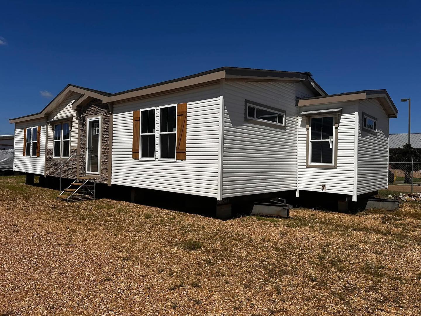 A white mobile home with brown shutters stands on gravel under a clear blue sky. It features a stone-accented entrance and multiple windows, conveying a rustic, serene atmosphere.