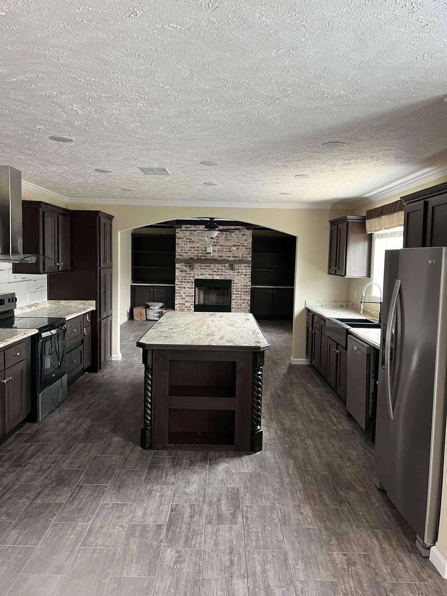 Spacious kitchen with dark cabinets, central island, and stainless steel appliances. Marble countertops add elegance. Brick fireplace in the background.
