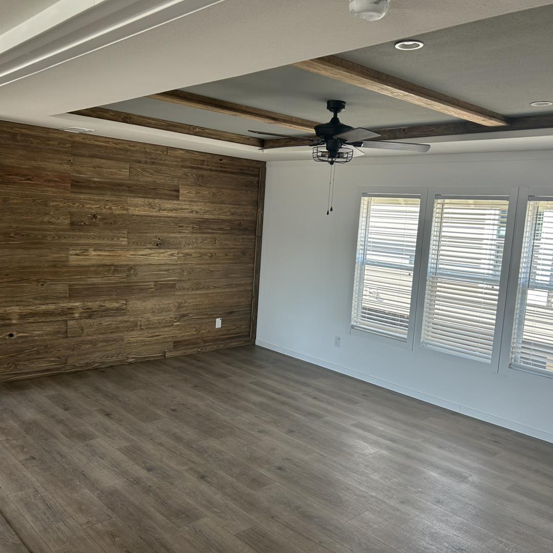 Spacious, empty room with wooden floor and accent wall, white ceiling with wood beams and fan, and three windows with blinds. Bright and modern.