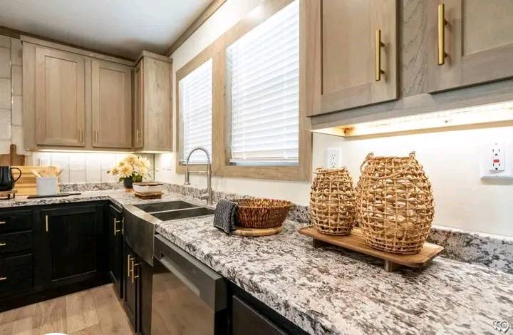 Modern kitchen with granite countertops, beige cabinets, and woven baskets. Sink under two large windows with blinds. Warm, cozy ambiance.