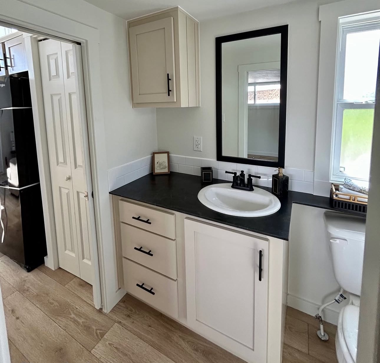 Compact bathroom with light beige cabinets, black countertop, and sink under a large mirror. Minimalist decor; wood floor adds warmth.