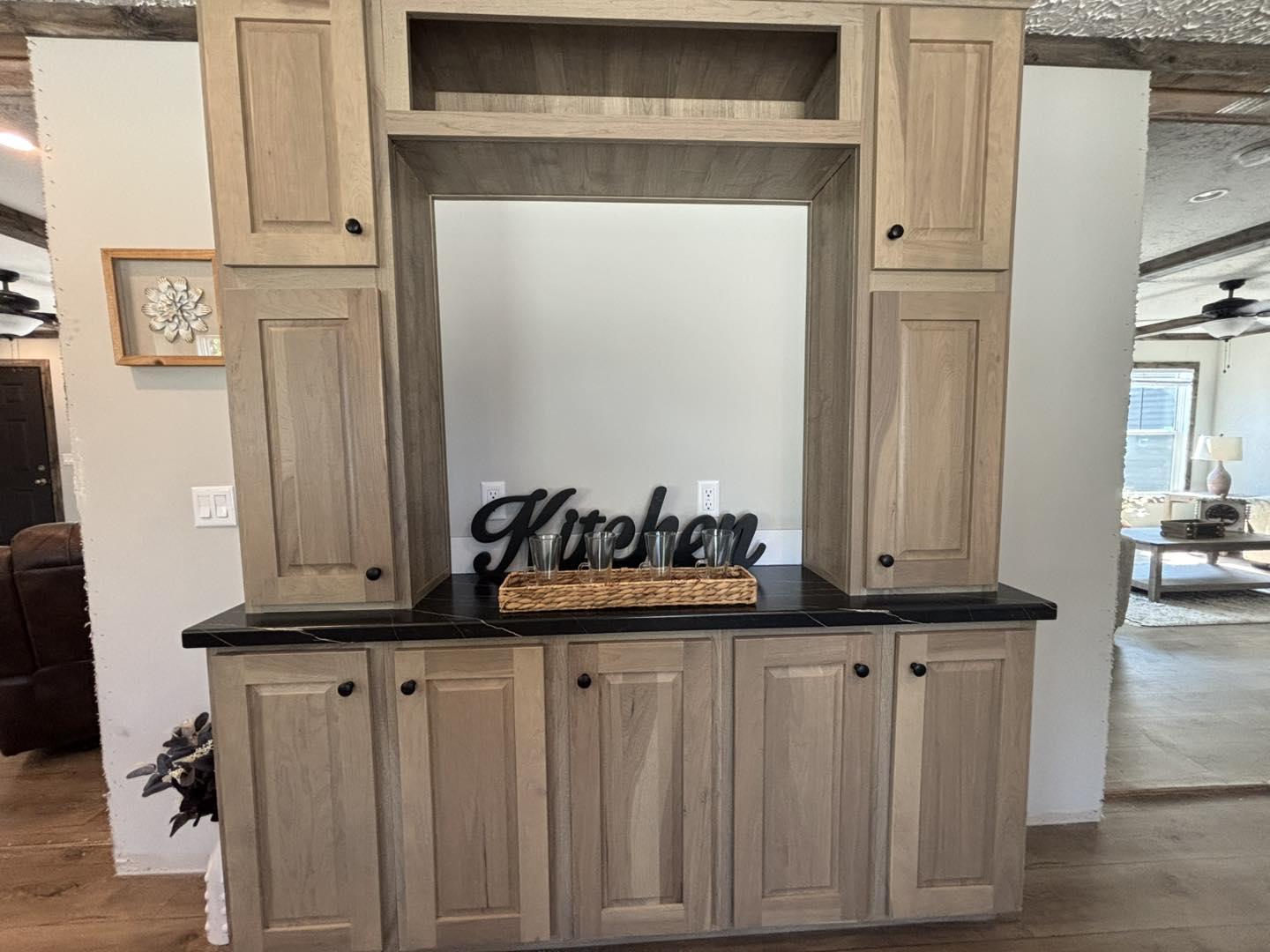 Wooden kitchen cabinets with a black countertop and "Kitchen" sign in the center. The room has a bright, cozy ambiance with visible living space.
