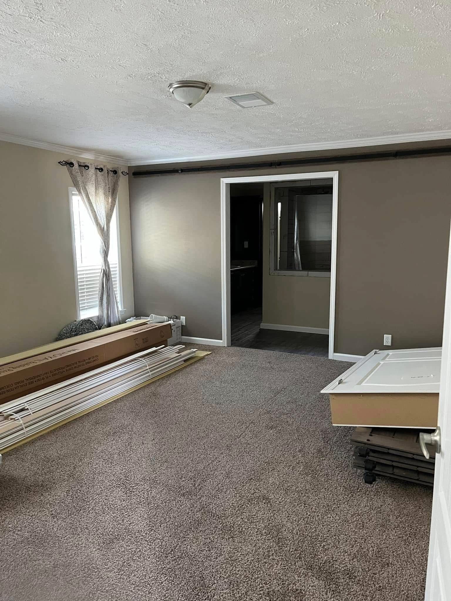 Empty room with beige walls and carpet, partially assembled furniture on the floor. Soft light enters through a window with sheer curtains, creating a calm, unfinished atmosphere.